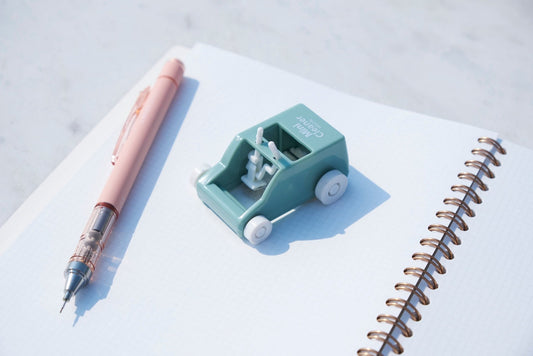 a Japanese toy stationery with wheels and a pink mechanical pencil sitting on top of a notebook under bright direct sunlight